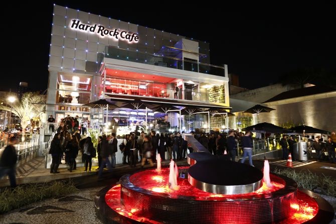 Fachada iluminada do Hard Rock Cafe Curitiba à noite, com multidão na entrada e o chafariz vermelho em primeiro plano