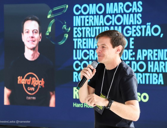 Rafael Magosso falando ao microfone no palco do FoodCo. Experience, com o telão ao fundo
